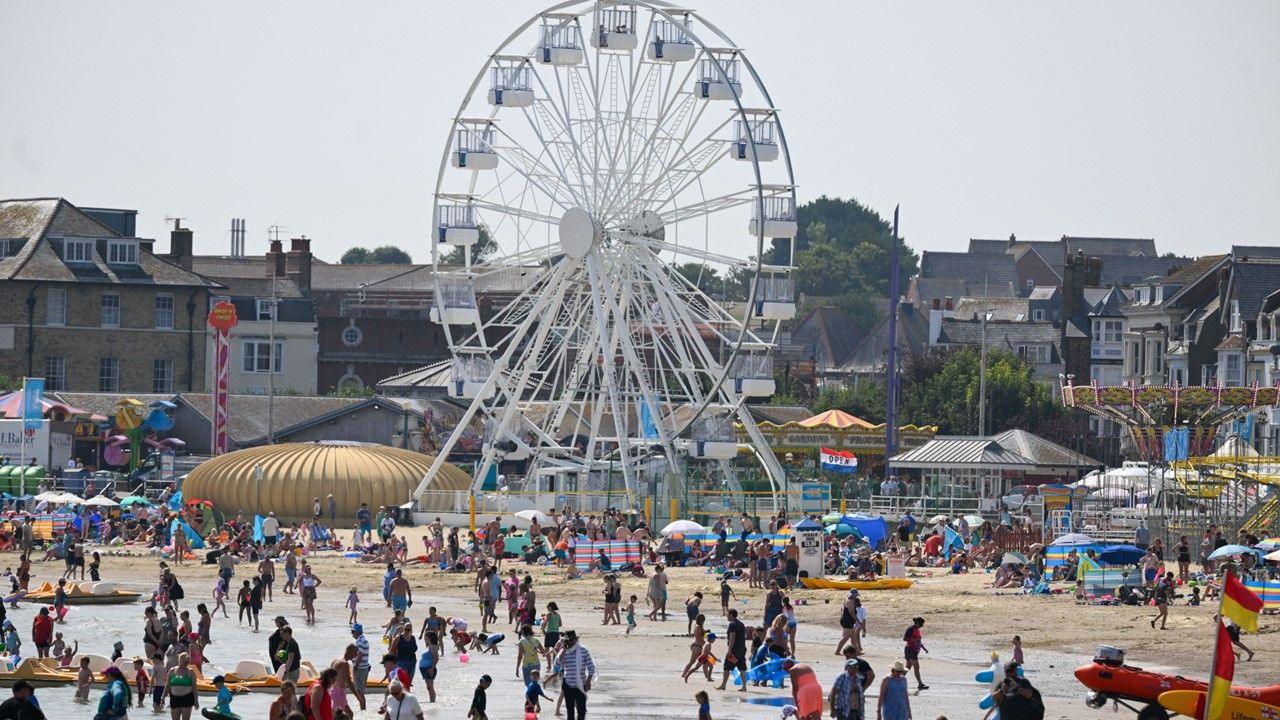 busy and colourful beach packed with families on sand with umbrellas and in shallow sea with pedalos, amusement park and big wheel behind, houses in distance, hazy coloured sky