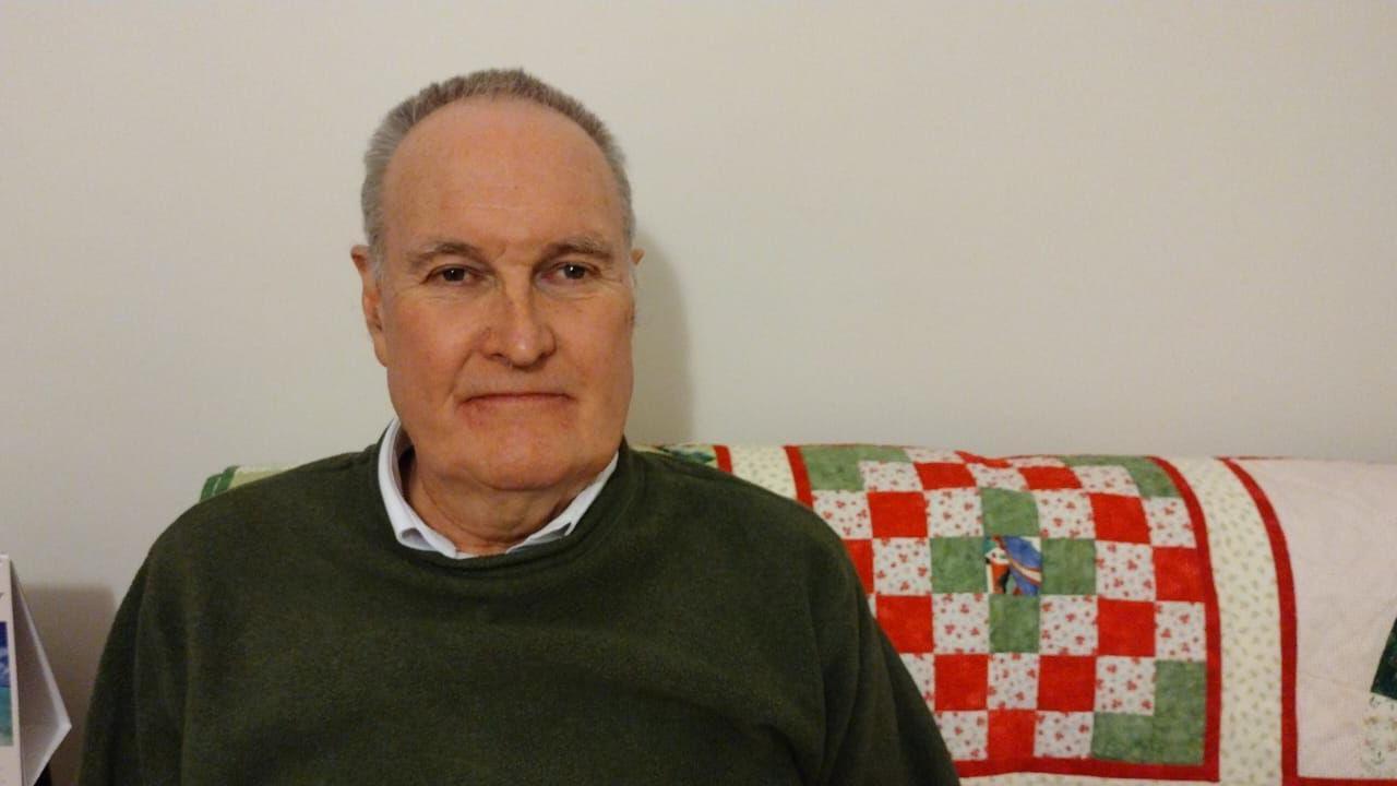 David Evans, with very short, grey hair, sitting on a sofa in a collared shirt and green jumper