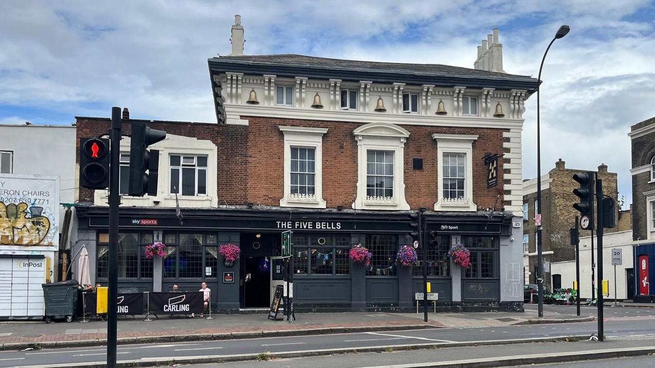 A large public house on a main road in London in three storey Victorian building. The ground floor exterior is painted a dark blue and there are flower baskets outside the pub.