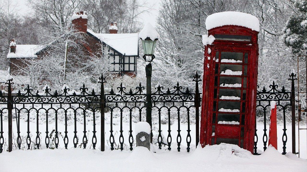 A bright red phone box next to railings topped with snow and snow covering the ground, house and trees behind