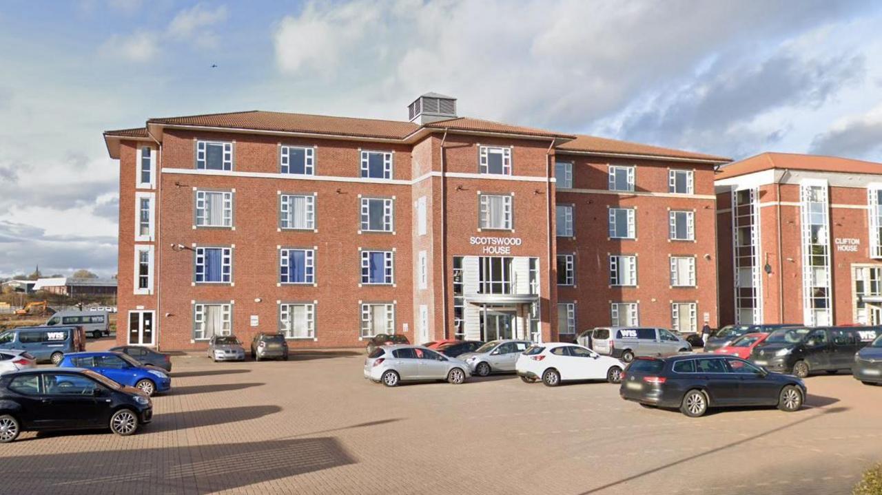 Scotswood House in Thornaby. It is a large four-storey building made from red bricks. Similar buildings stand on either side of it and a large car park is located in front of the main entrance.