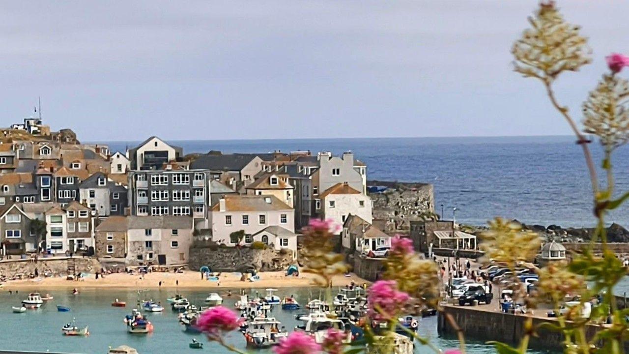Houses and buildings with a beach in front surround a bay with boats. Blue sea and hazy sky behind 
