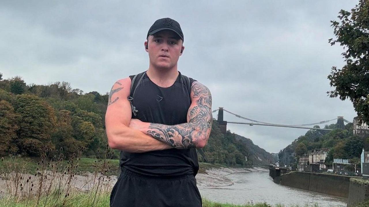 Ashley stands with his arms folded in black running gear, including a black cap. He has a tattoo sleeve on his left arm. Behind him the Bristol suspension bridge, River Avon and a road running along it can be seen.