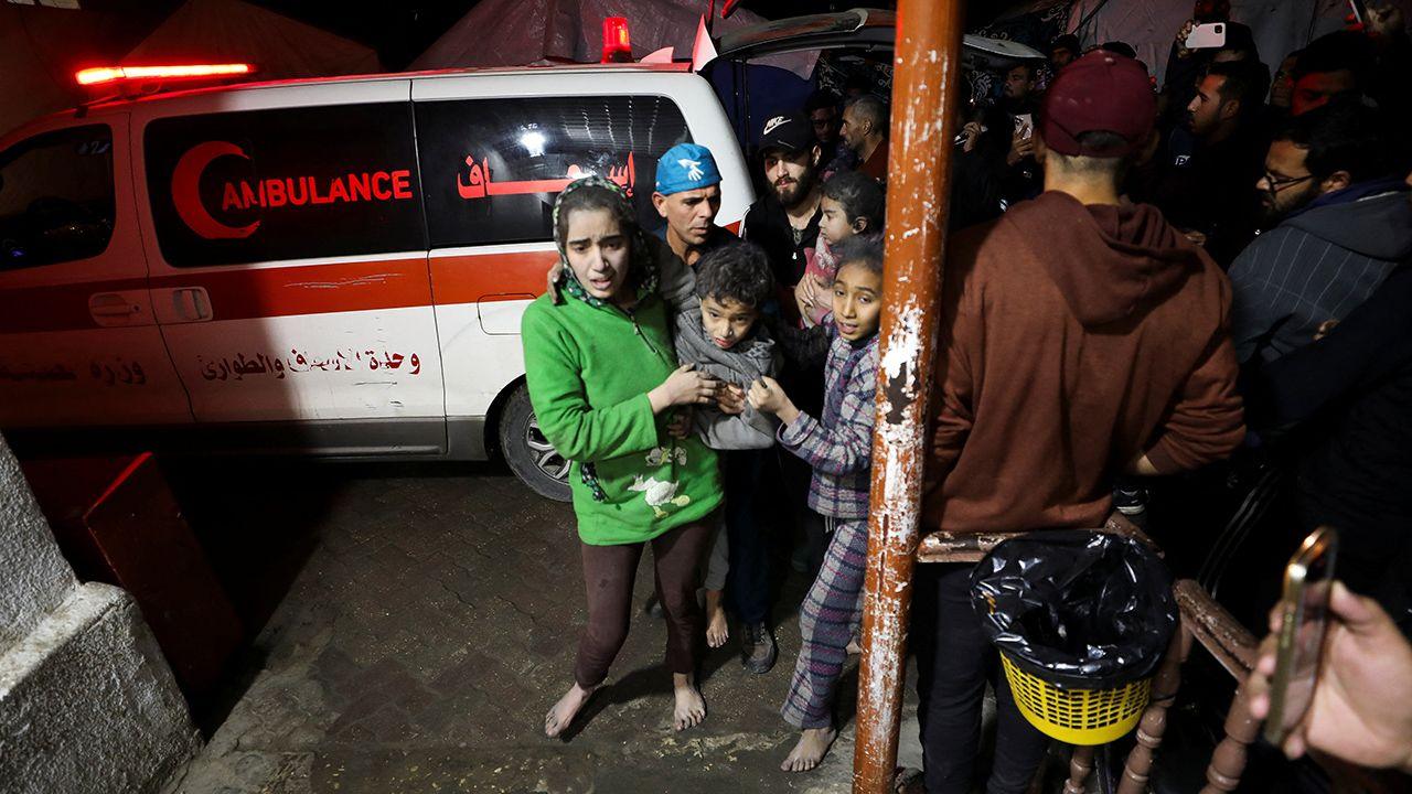 A group of Gazans stand next to an ambulance, looking distressed. One woman wears a green top and carries a child.