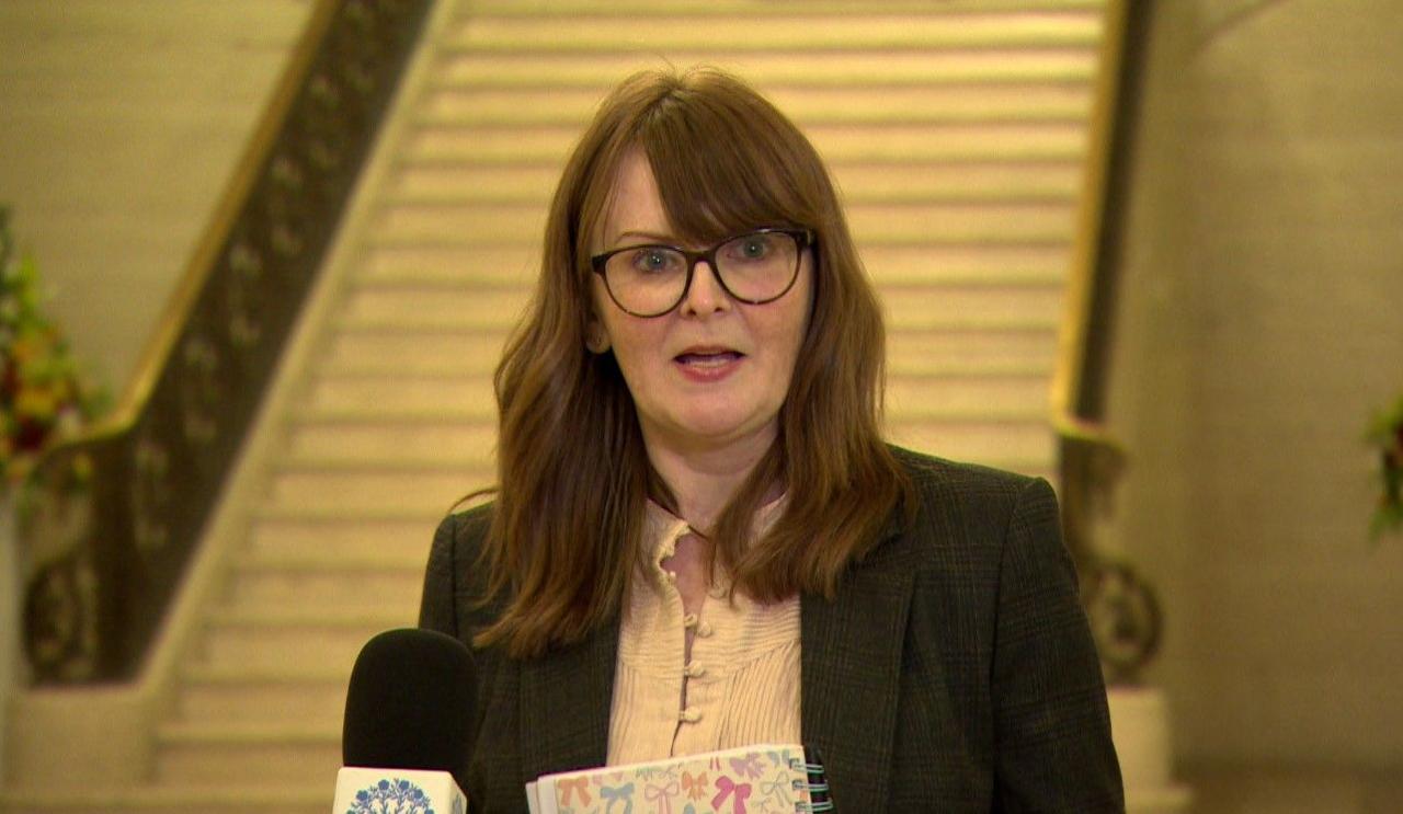 Caoimhe Archibald has mid length brown wavy hair. She wears brown rimmed glasses, has red lipstick and wears a white shirt and a black jacket. She is mid sentence in this screengrab from the Great Hall in Stormont, taken at about 17:00.