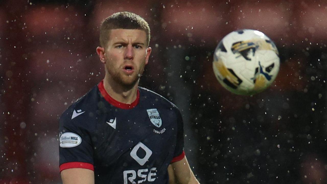 Ryan Leak in action for Ross County. He is wearing a dark blue shirt and is watching the ball move away from him after sending a long pass forward.