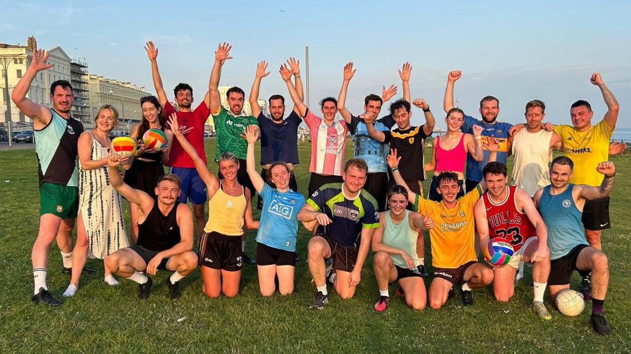Members of Brighton & Crawley Gaels Gaelic football club pose for a photo on Hove Lawns by the seafront. They are wearing an assortment of colourful t-shirts and are cheering with raised hands. The sky is clear blue, with the i360 visible in the background.