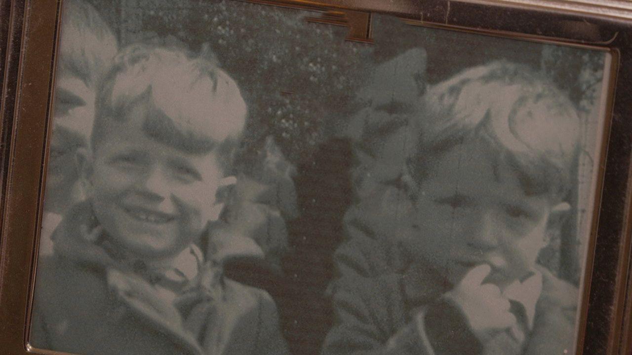 Sepia-coloured photo of two young boys with pale coloured hair. One smiles while the other looks pensive and has a finger in his mouth.