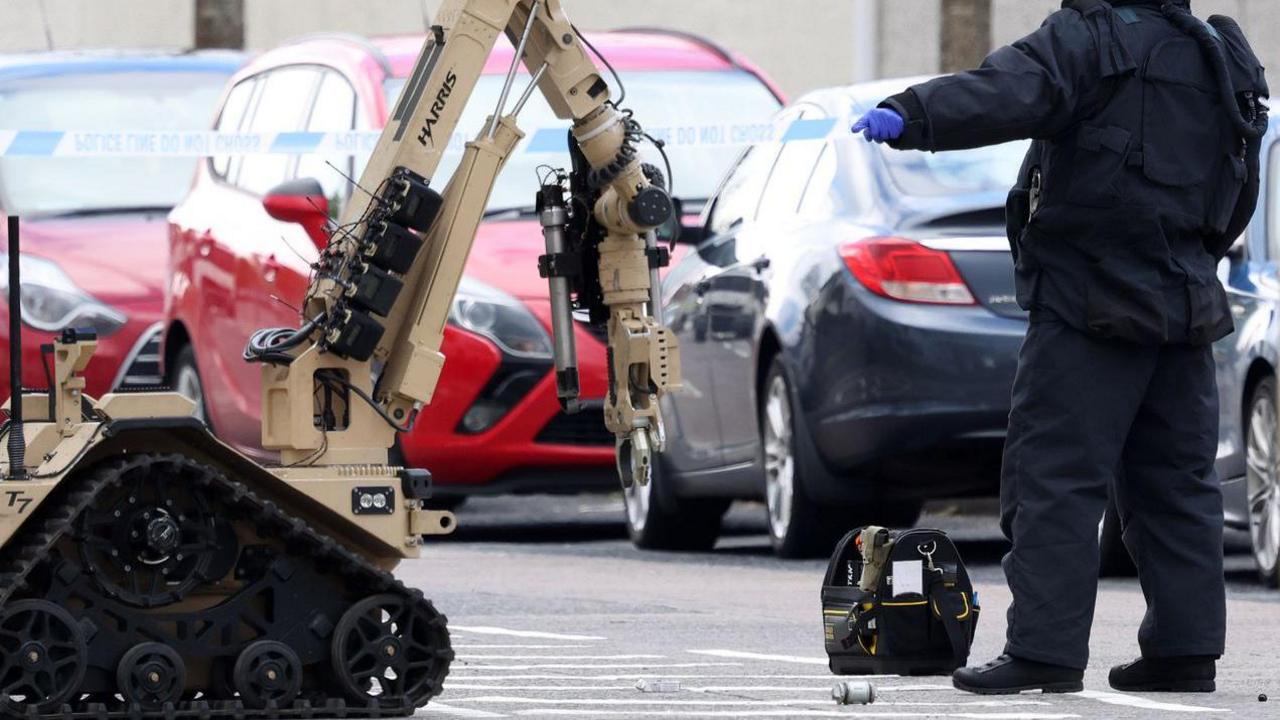 A bomb disposal officer in navy overalls and a black helmet, stands with his back to the camera. To his left is a sand coloured robot with four black wheels and a claw. On the floor is a small steel pipe. Cars can be seen in the background of the frame.