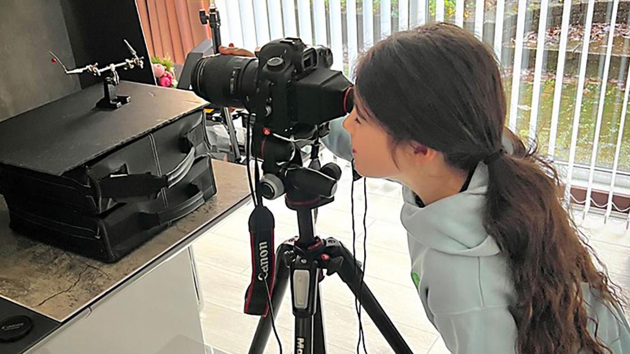A girl with long brown tied-back hair, wearing a light blue hoodie and looking through a black camera on a tripod.