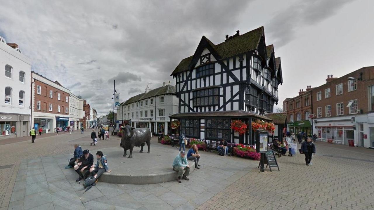 The Black and White House Museum in Hereford. The building has Tudor-style black beams and flowers hanging in baskets around the front. It stands in a pedestrianised square with people sitting on a pedestal in front of it