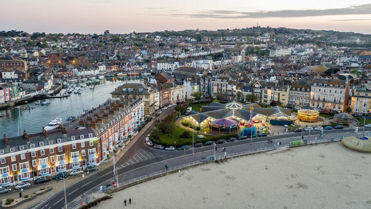 Weymouth's promenade as seen from above. A sandy beach has a wide footpath and a road running along it. Rows of houses are set back from the beach as well as a number of carousel rides.