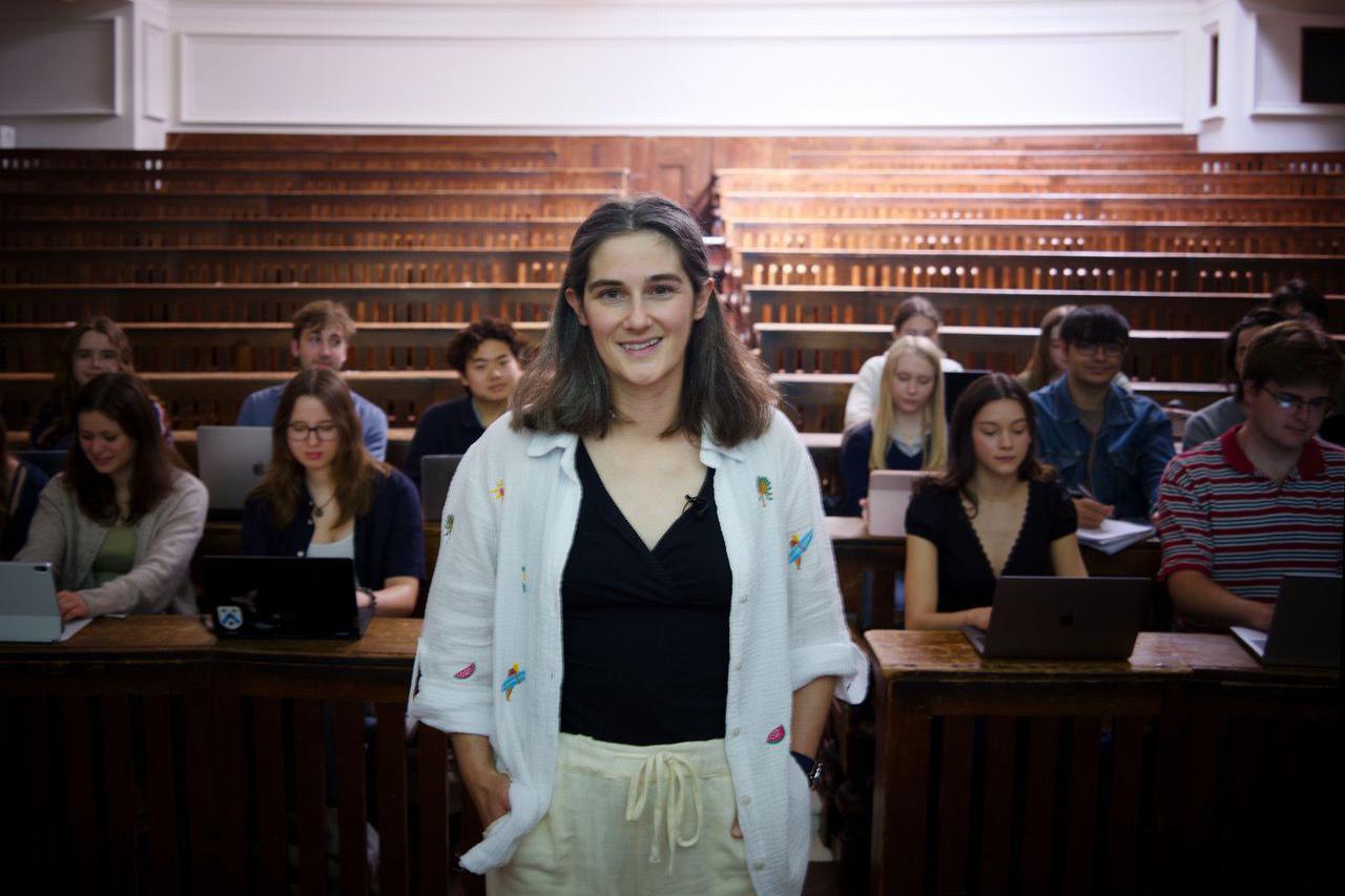 Dr Cole stands at the front of a wood‑panelled lecture theatre, facing the camera, with several rows of tiered seating behind them. Other people sit at desks in the background using laptops and tablets, suggesting an academic setting. The room features warm lighting, wooden benches, and a symmetrical layout.