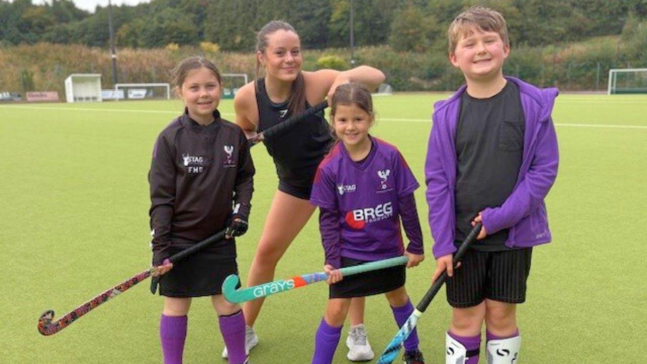 Two small girls and a boy, dressed in black and purple hockey kits, hold hockey sticks up as they smile at the camera. A woman dressed in a black sports vest and shorts, stands behind them and smiles as she holds her hockey stick up high.