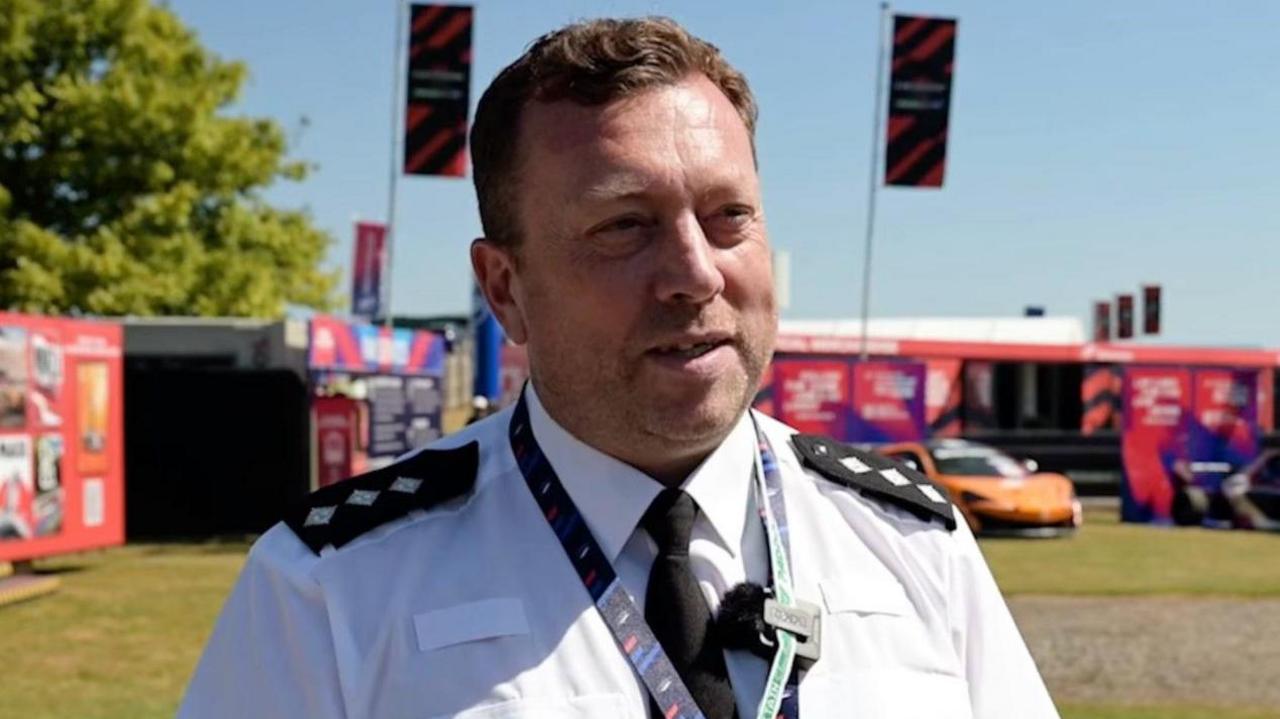 A police officer in a white shirt and black tie. He is standing within the grounds of a motorsport track.