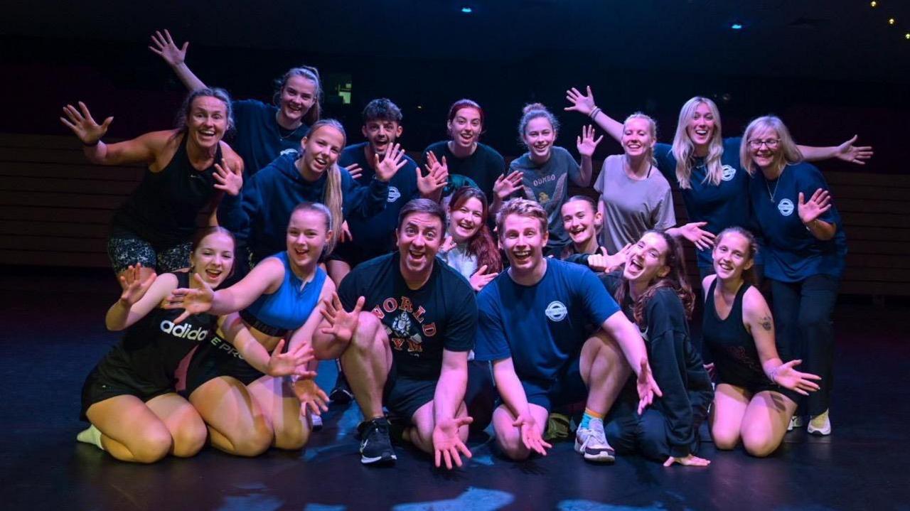 A group of dancers standing and crouching in a group. All of the people in the picture are wearing sports clothes, smiling at the camera and waving their hands. 