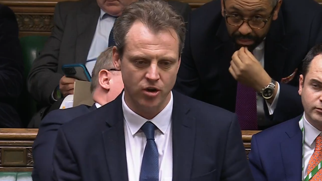 Joe Robertson is wearing a dark blue suit and blue tie and is talking to the House of Commons.