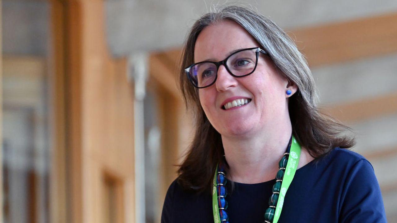 Maree Todd walking in the Scottish Parliament. She is wearing glasses and is wearing a blue top, a blue and green necklace, and a green lanyard