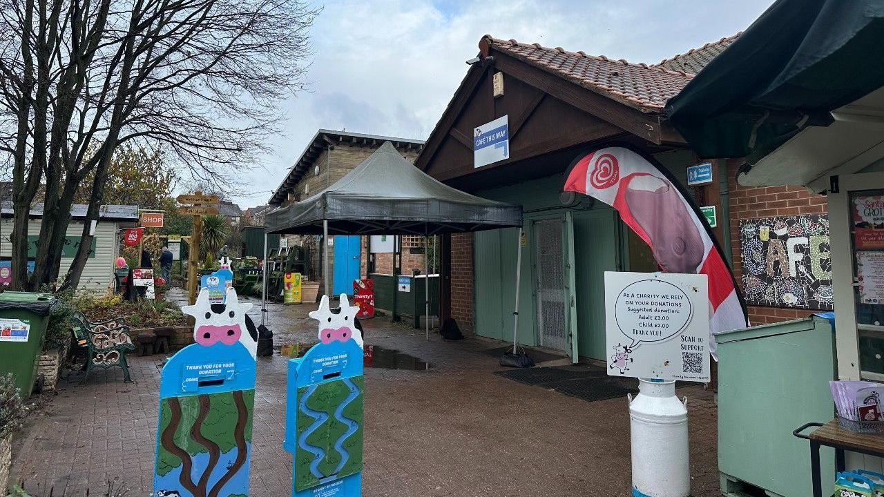 The reception area of Stonebridge city farm, showing a series of single storey buildings with cow-themed signs outside, linked by a gravel path with gardens visible in the distance