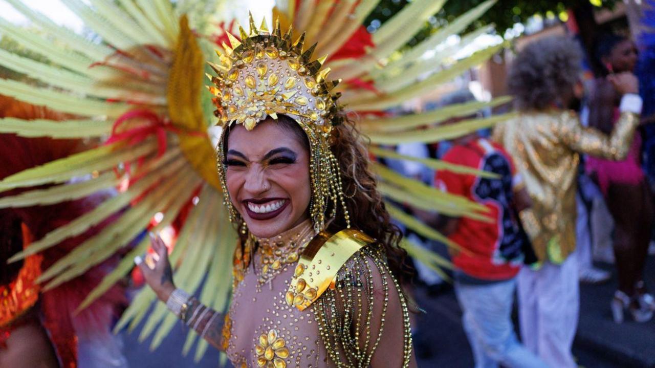 A woman in a gold headpiece, golden body jewels and performance makeup smiles at the camera.