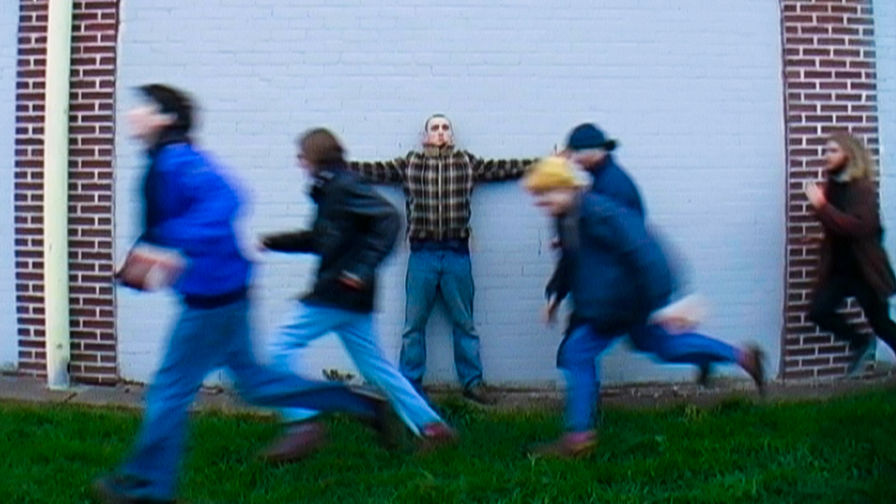 Picture is an album cover showing a man standing in front of a white brick wall and five men running past him.