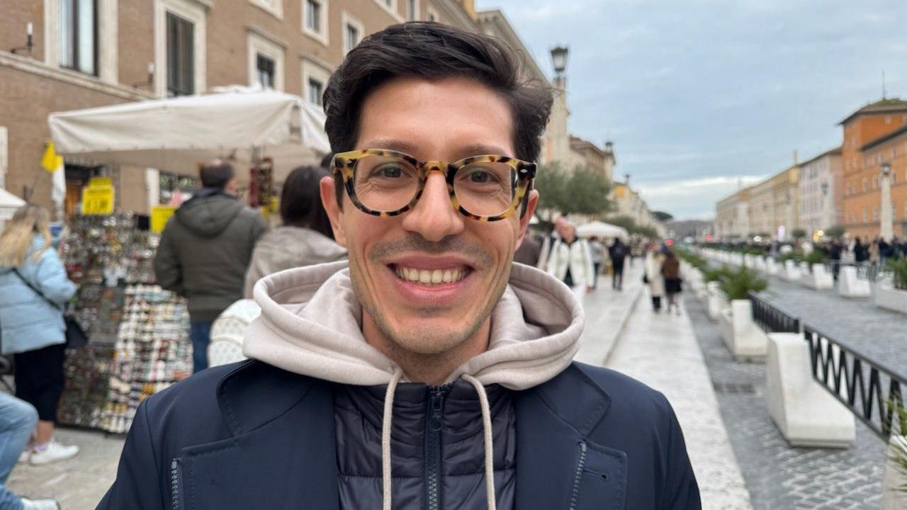 Man with dark hair and glasses, wearing a jacket and hoodie and smiling outside of St Peter's, Rome