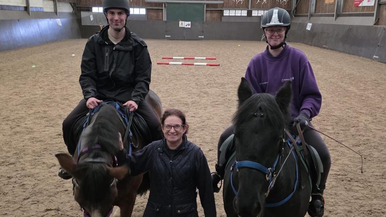 Two riders are sitting on top of black horses with a woman stroking the neck of the horse on the left. There are some red and white striped poles lying in the paddock behind them.