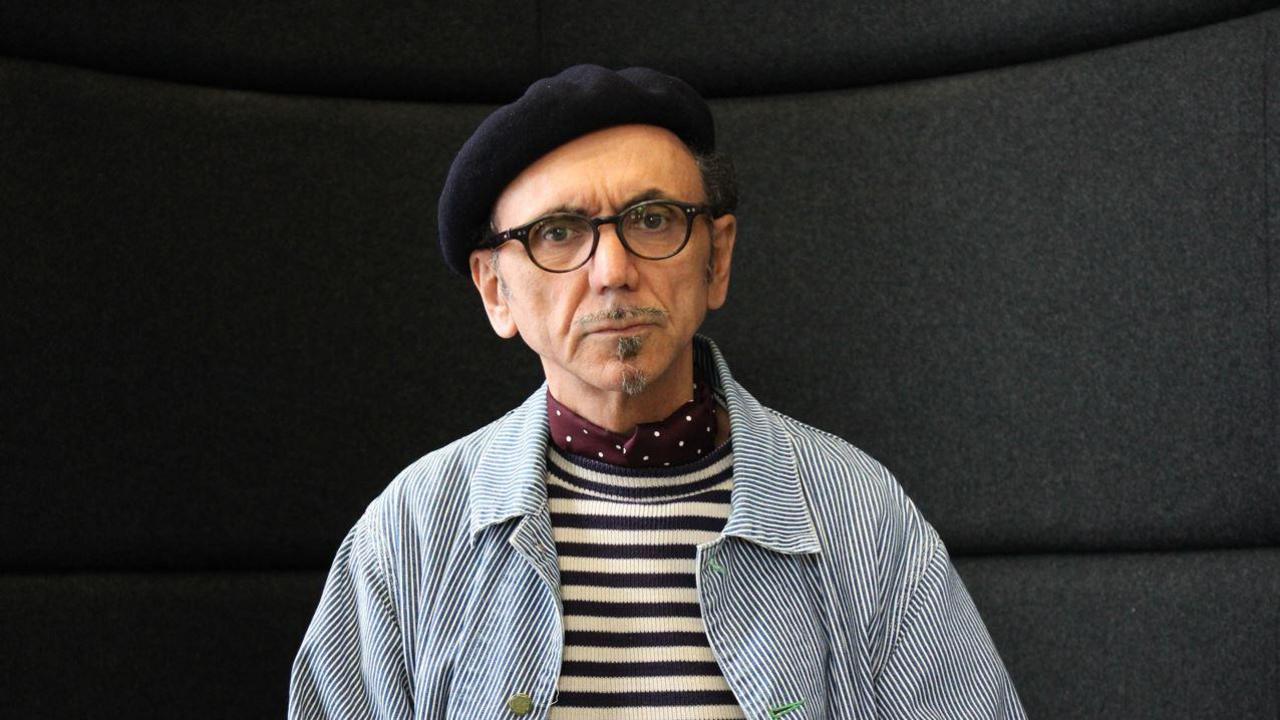 A man with dark glasses, a small goatee beard a black beret and serious expression in a blue and white striped jacket and black and white striped tshirt in front of a black background