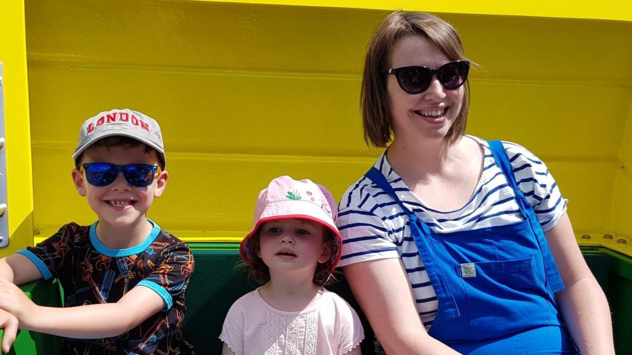 Nicola with Oscar and Ffion. The children are wearing sunhats and t-shirts and Nicola is wearing blue dungarees over a blue and white striped t-shirt. All are smiling. 