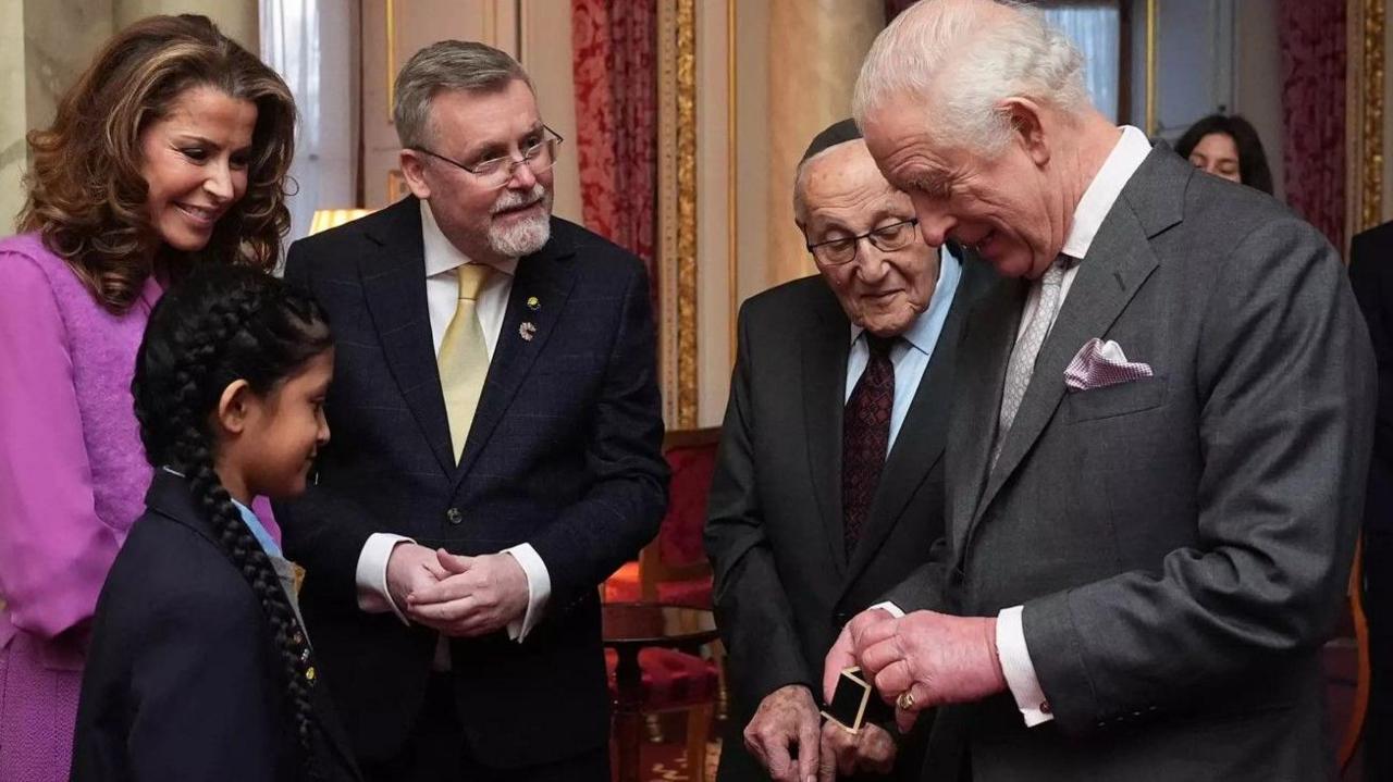 Left to right, those photographed are Natasha Kaplinsky smiling in purple, child Yaashvi, Adrian Packer, Manfred Goldberg. The two men are wearing glasses, a dark jacket and a tie. King Charles is holding a tin ring.