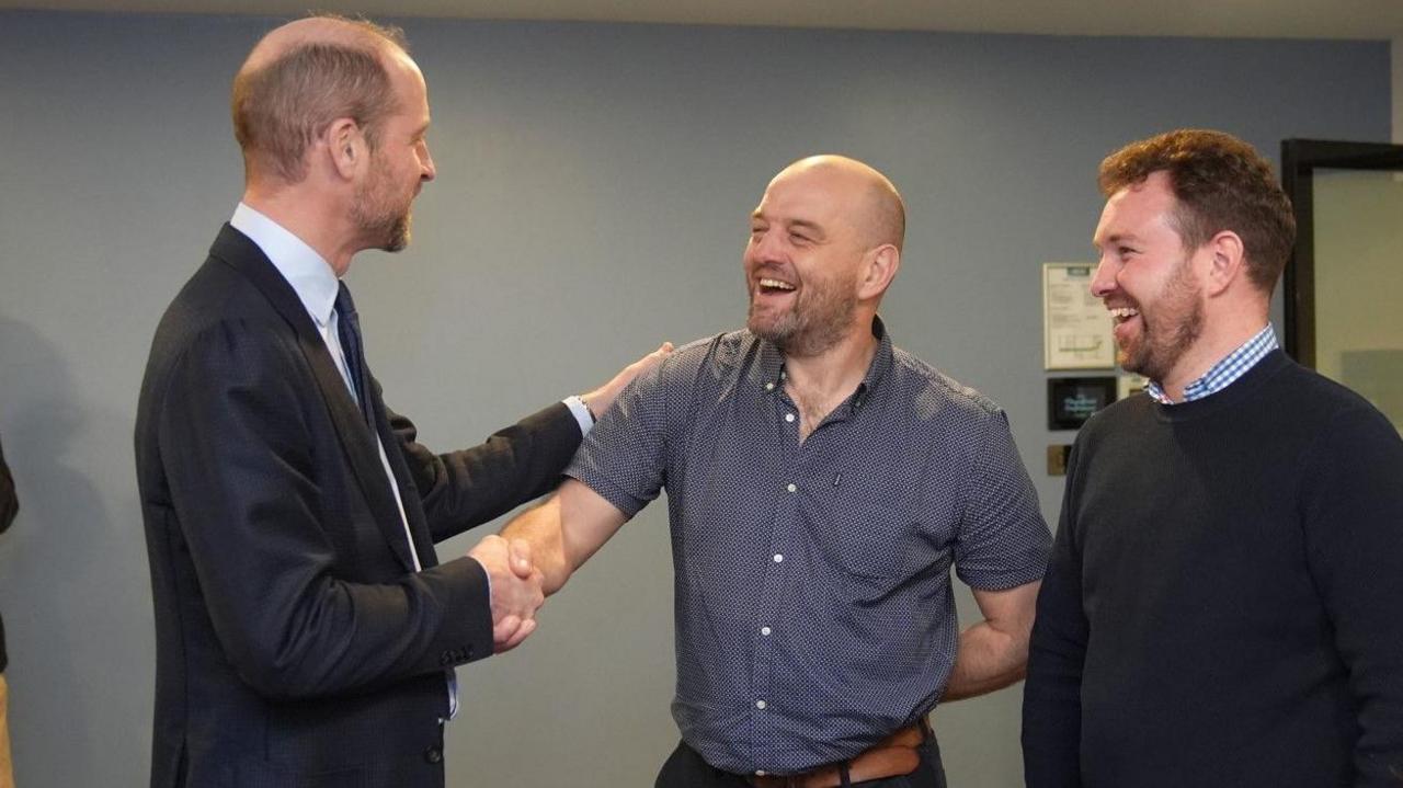 The Prince of Wales is shaking hands with and touching the shoulder of paramedic Ed Griffiths. Another man is smiling on the right of the photo.