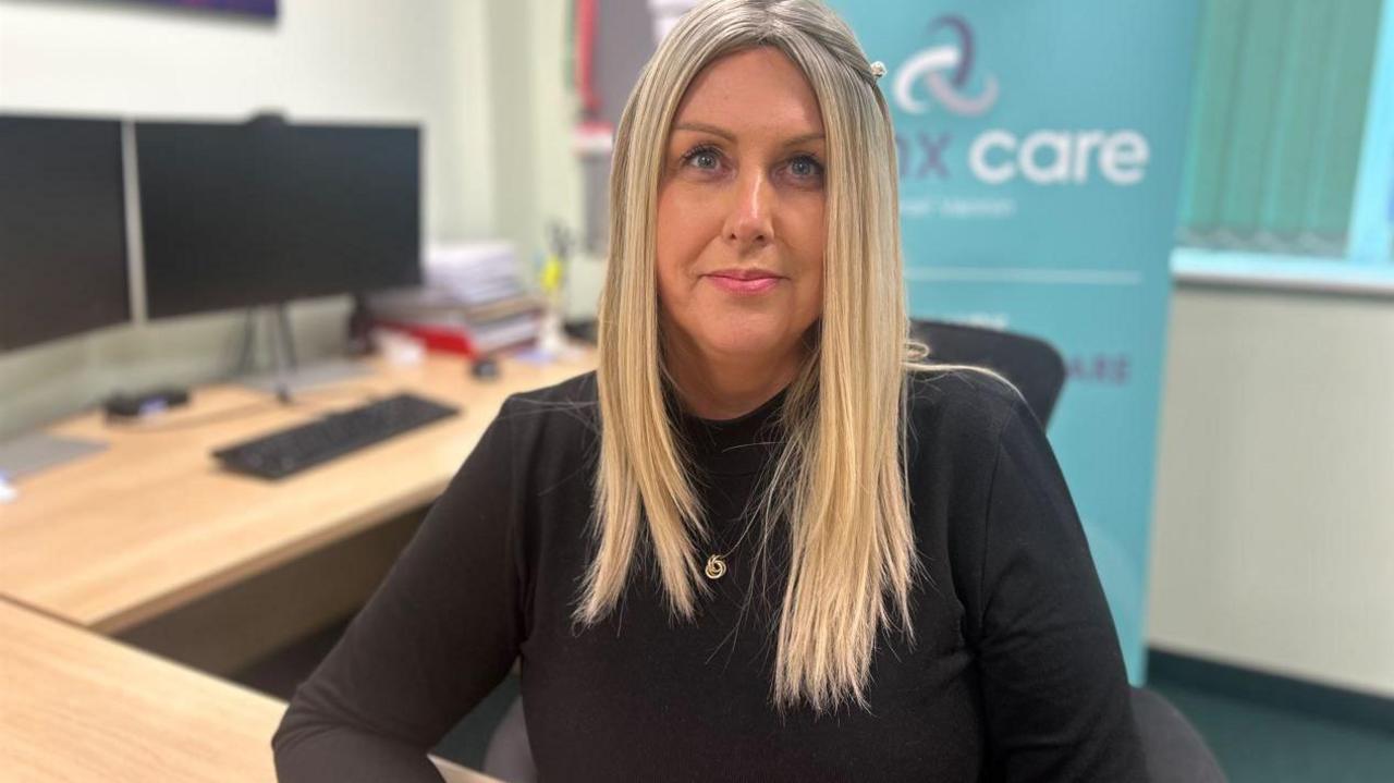 Teresa Cope, a woman with blonde hair wearing a black long-sleeved top. She is sitting at a desk with a Manx Care sign in the background. 