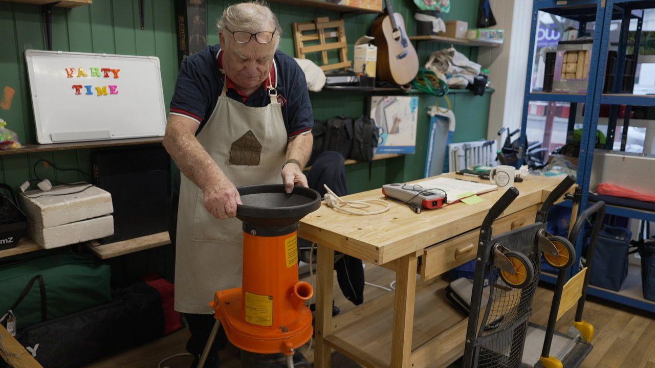 A 74-year-old man is wearing a blue and red polo shirt and a pale blue/grey  apron with a brown shed on the front. He's looking down at an orange power tool which comes up to his hip and looks like a leaf shredder. To his left is his work bench which is covered in electrical testing equipment. Behind that is a row of shelves holding random objects such as bunting and guitars. Over his right shoulder is a white board and magnets have been arranged on it to say 'party time'.