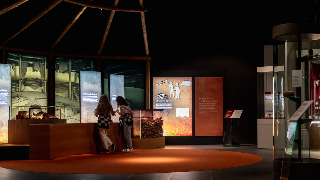 A dark room with an circle orange carpet and screens along the walls. There are two young girls looking inside a glass casing - situated in front of the screens. It is part of the new gallery. 