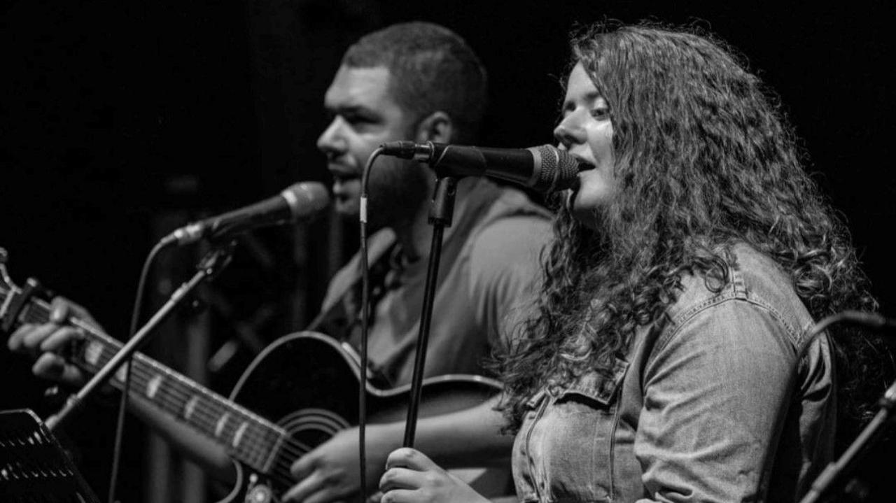 A black and white photo of Andrew and Georgia singing into microphones. Andrew is playing an acoustic guitar.