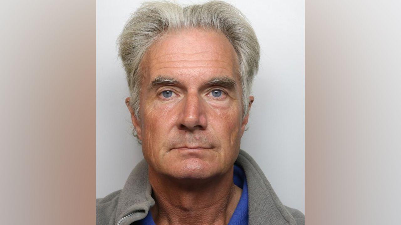 A a mug shot showing an older man with grey hair and eyebrows. He has blue eyes. He is wearing a grey fleece.