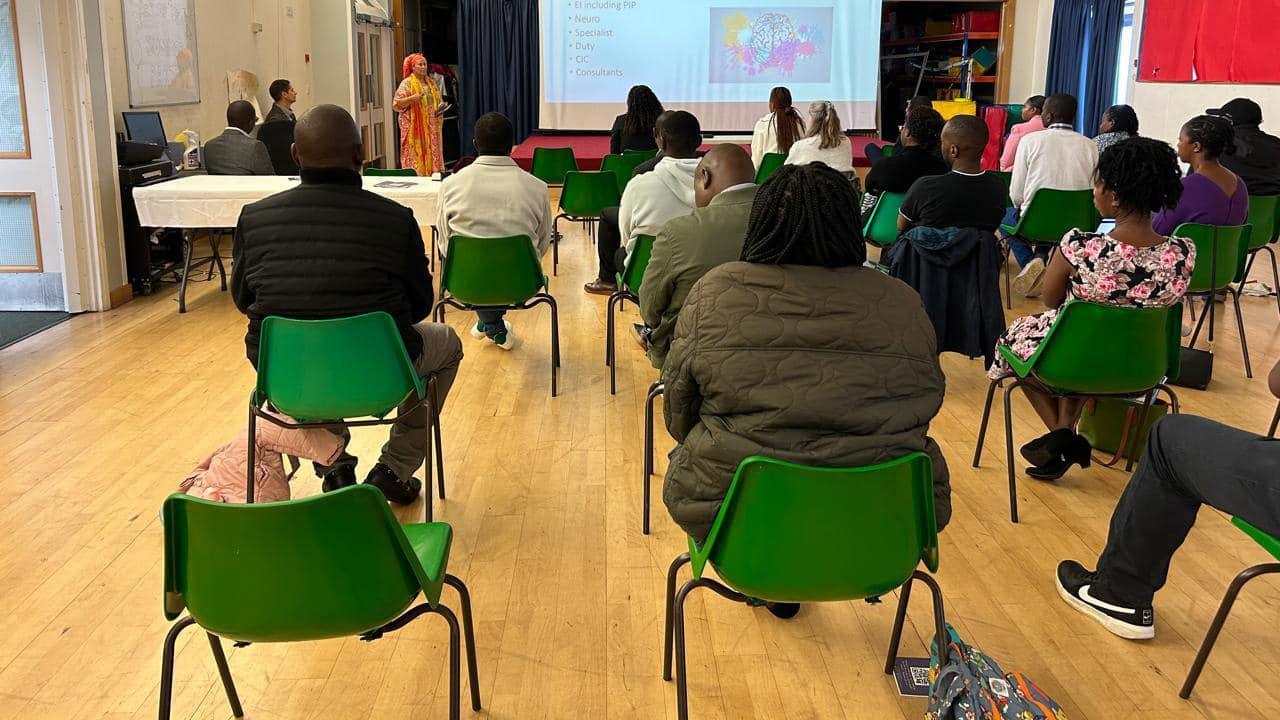 Dozens of people sit on green plastic chairs as they look at a woman who is giving a speech next to a projector screen in the background. She is wearing a brightly coloured dress.