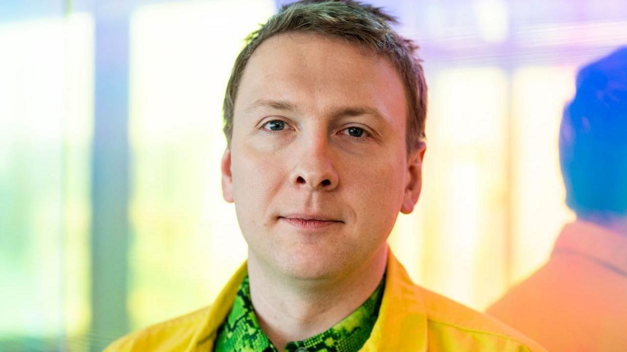A head and shoulders shot of a man in a yellow jacket and green snakeskin shirt. He is looking at the camera and standing with his back angled to a mirror so his reflection can be seen to the right. Glass behind him has a yellow and green hue. 