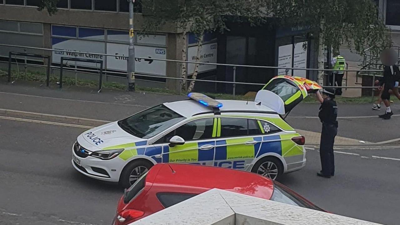 A female police officer closes the boot of a police car that is parked in the middle of a road to block traffic, with two teenage boys walking on the pavement behind her.