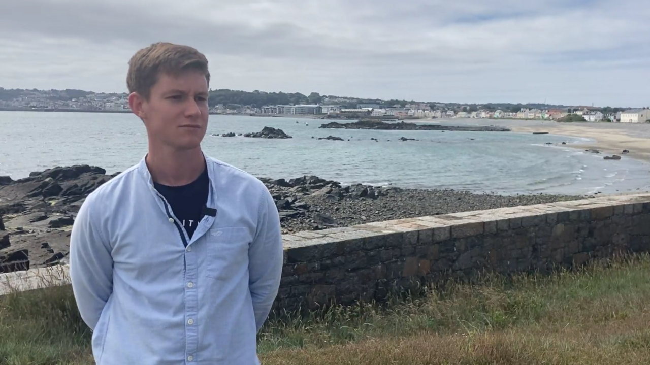 Brandon Ashplant, standing near a rocky shoreline with the sea in the background. He has blonde hair and is wearing a light blue button-up shirt over a dark-coloured shirt.