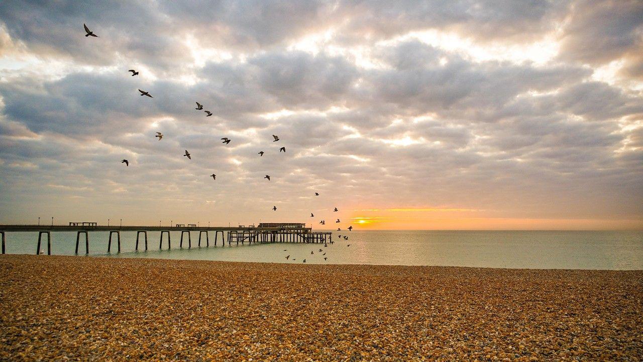 Birds fly above a shingle beach and pier under a cloudy sky, with a low Sun just visible through the clouds
