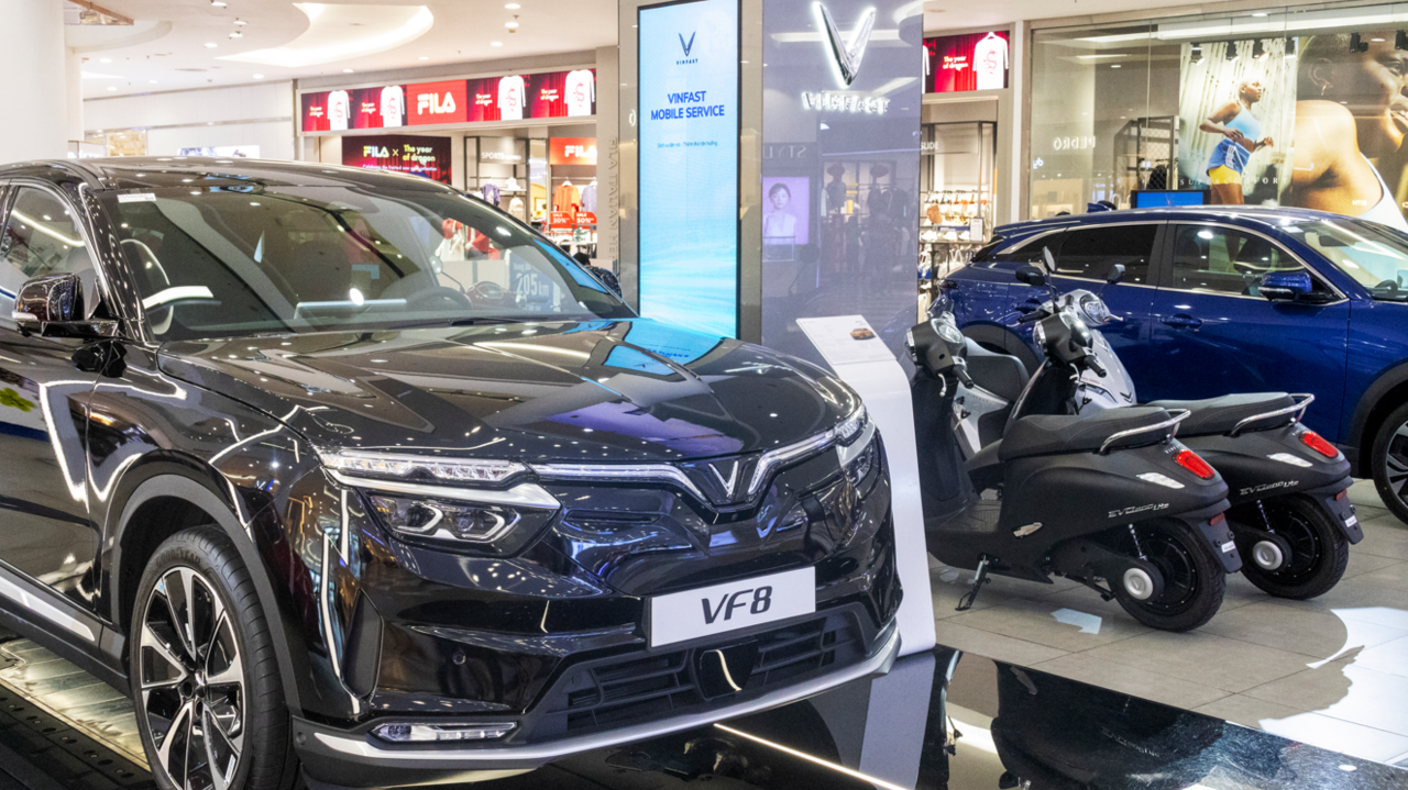 A black car in a showroom with the letters VF8 on the number plate and two black scooters and a blue car in the background