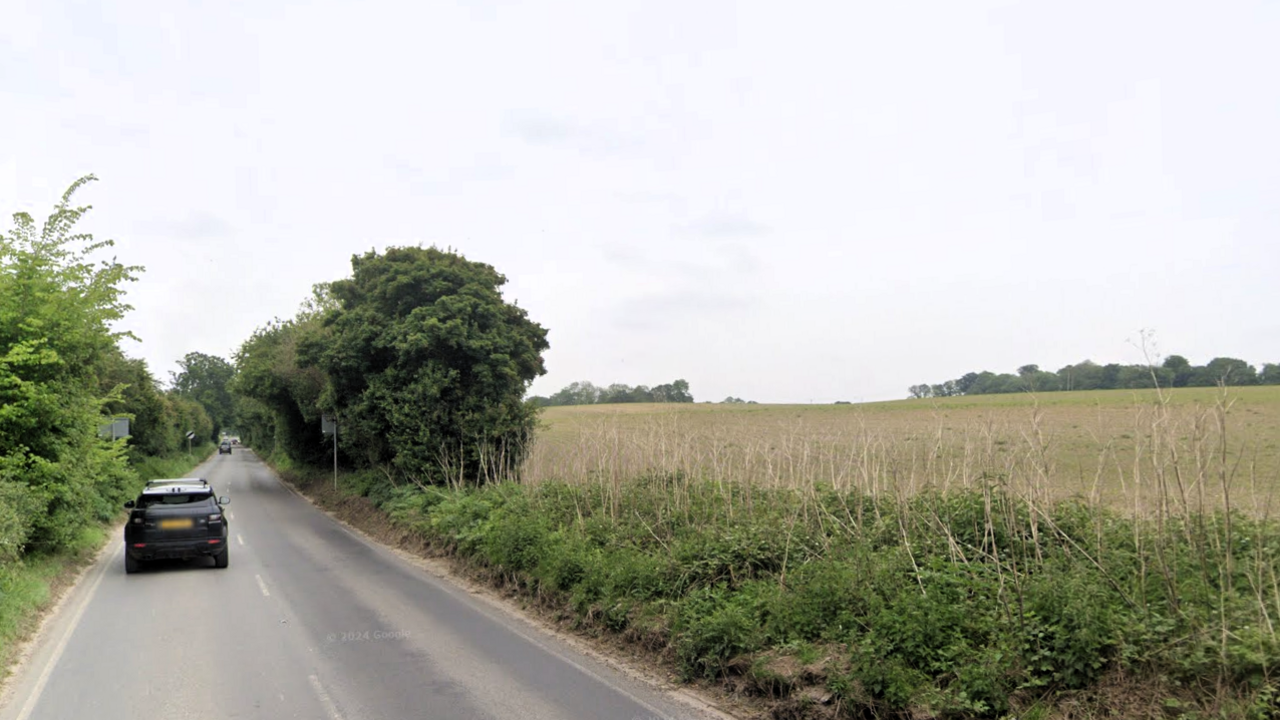A car travels along a tree-lined road to the left of the picture, with a harvested field to the right and trees on the horizon.