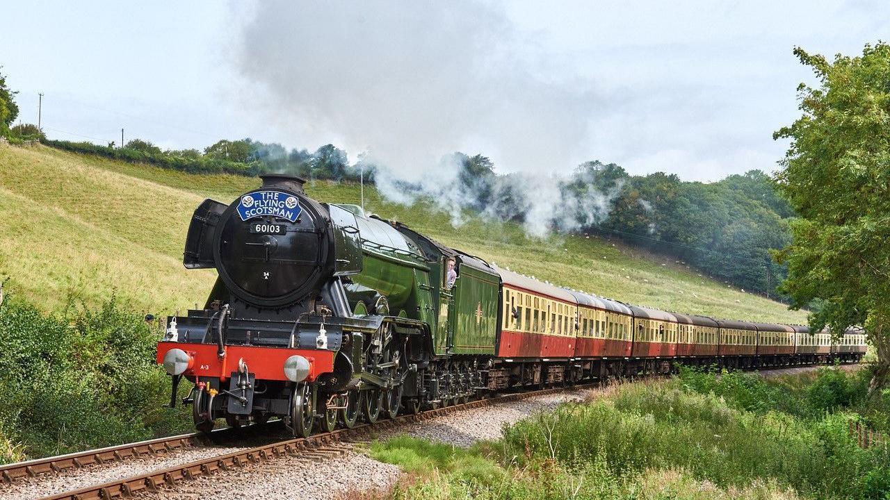 The historic Flying Scotsman train is going along a track with a hill to one side. Steam is rising from the engine.