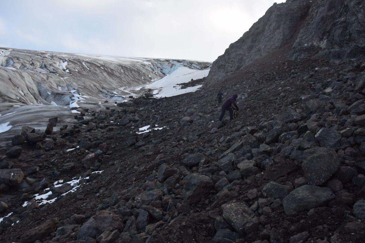 A large plain of black rock, with a valley behind with the glacier. A person stands on the rocks.