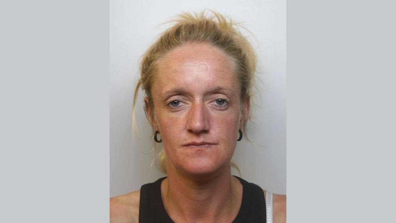 A police custody image of Lauren Thomas. She has long bleach-blonde hair which is tied back in a ponytail. She is wearing a black tank top and has black hoop earrings.