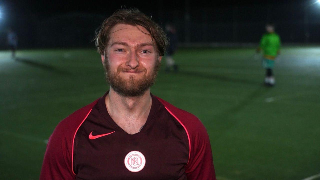 Lewis has dark, sweat-soaked hair and a ginger beard. He is wearing a red football shirt and standing on a green football pitch.