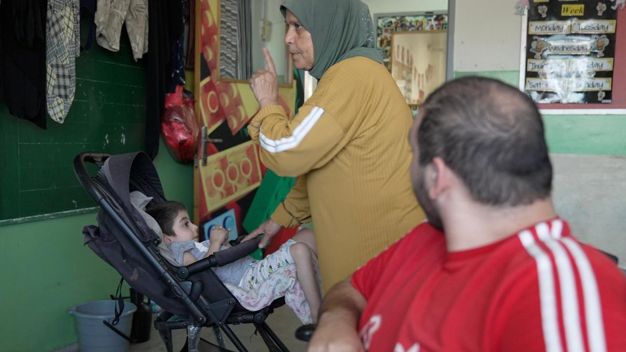 An older woman with a yellow headscarf has one hand on a pram trying to rock it, while inside is a slender young boy with brown close-cropped hair, wearing a grey T-shirt and pyjamma bottoms. Her other hand raises her finger to her lips to make a shhhhh sound to soothe him.