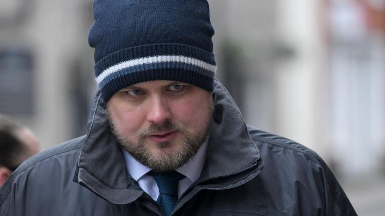 Ryan Mason wearing a white shirt and blue tie underneath a dark grey raincoat. He is wearing a dark blue beanie hat whit a white stripe, and is looking just off to the left of the camera with a stern expression.