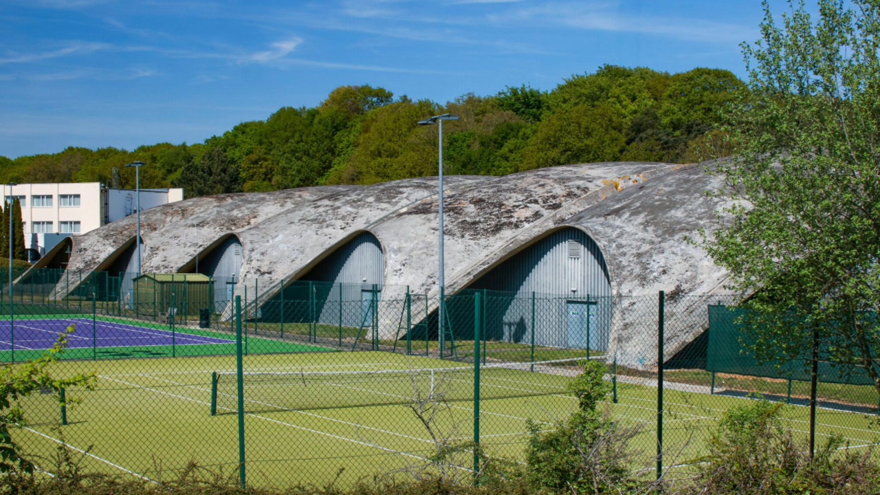 A row of shell-like concrete structures forming a roof to an oblong building. The concrete is grey and patched with black and yellow lichen. In the foreground are two tennis courts, one with a green playing surface, the other purple.